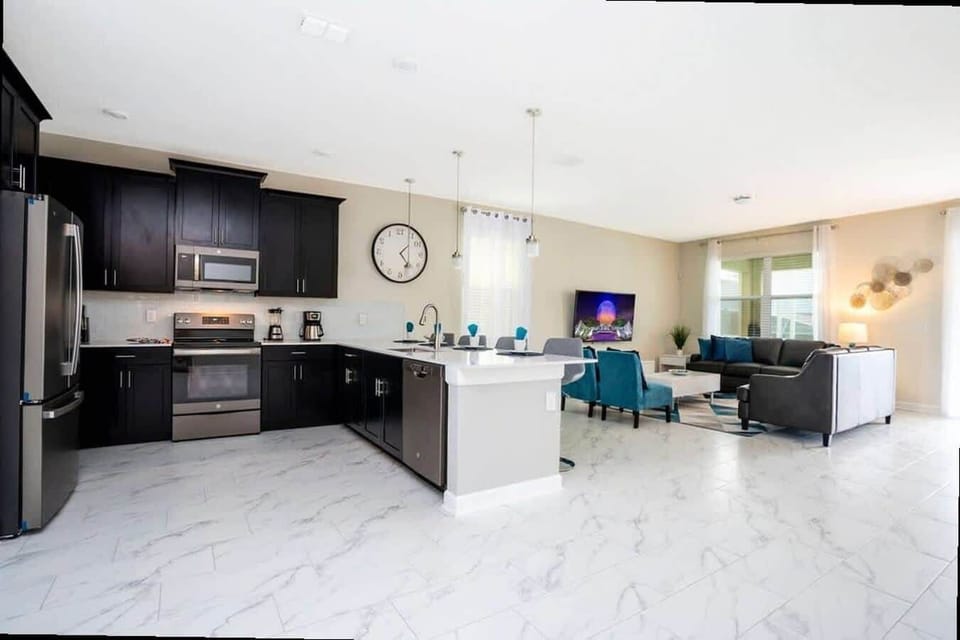 Enjoy open-plan living in this airy space: a sleek kitchen with dark wood cabinets, stainless steel appliances & quartz island for four flows into a cozy living room with gray sofa, teal accent chairs, modern wall art & bright light.