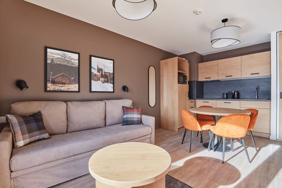 Living and dining area with a grey sofa, round table, orange chairs, and a kitchenette with light wood cabinetry and dark countertop.