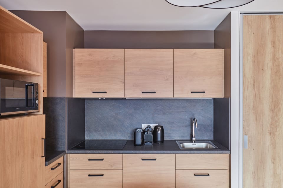 Kitchenette close-up with light wood cabinets, microwave, coffee maker, kettle, and a dark slate back panel — fully equipped.