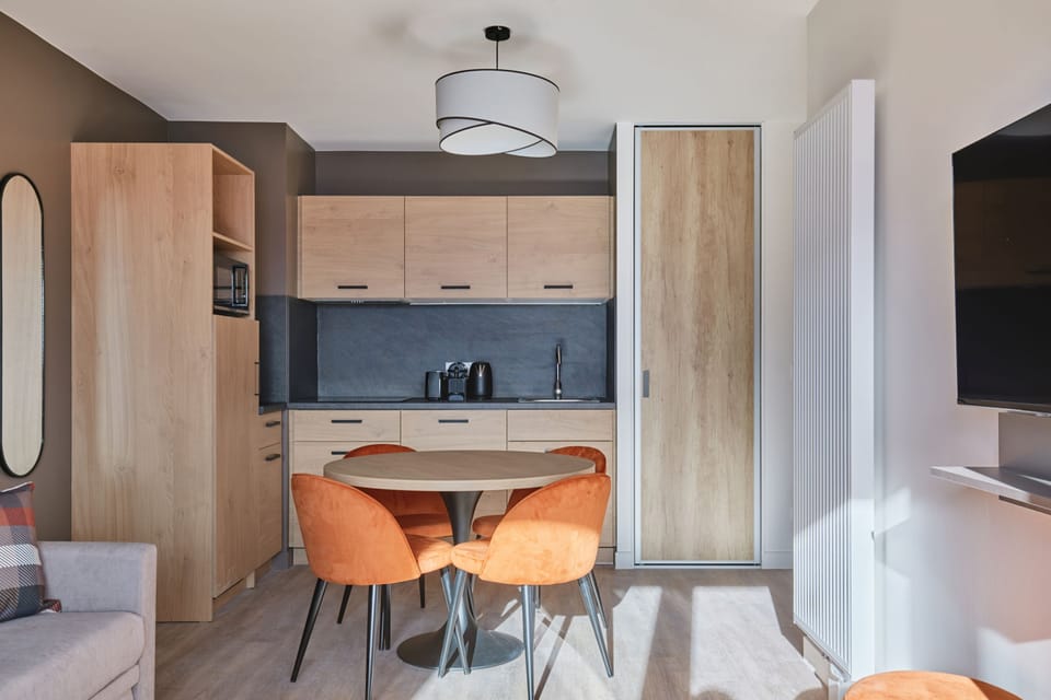 Dining area with round table and orange chairs beside a sliding door, with a wall-mounted TV visible in the background.