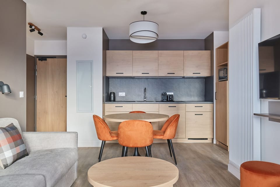Kitchenette with light cabinets, dark countertop, coffee maker, and kettle — the sofa with plaid cushions is visible in the background.