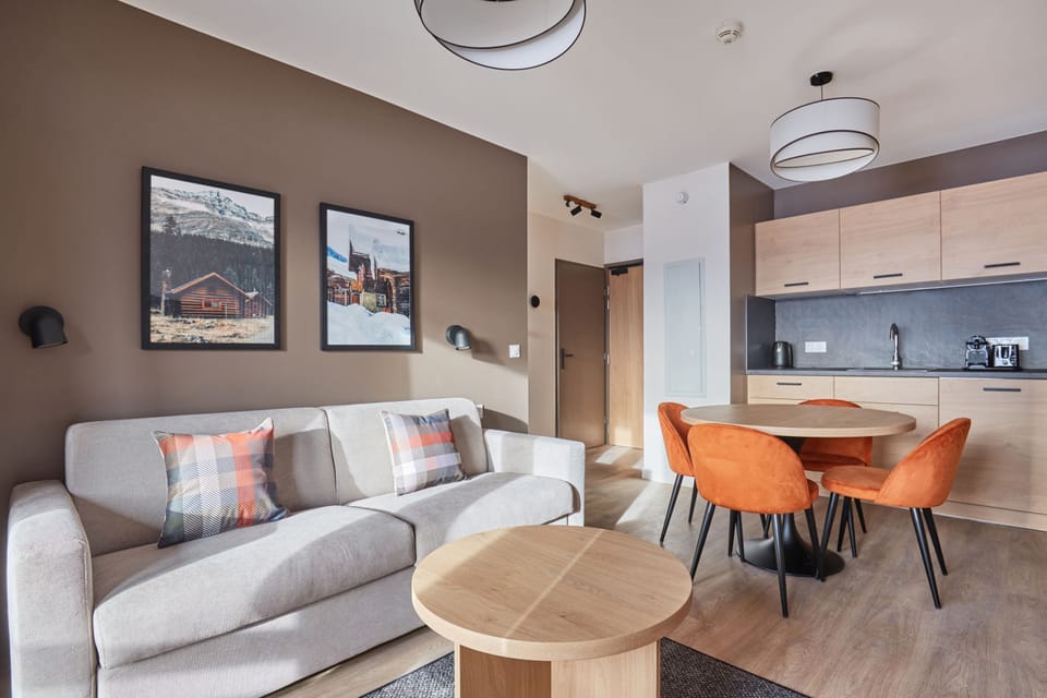 Open-plan living and dining area with grey sofa, round table, orange chairs, and a kitchenette with light wood cabinetry.