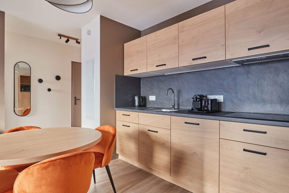 Kitchenette close-up with light wood cabinetry, dark stone countertop, sink, and orange dining chairs beside the round table.