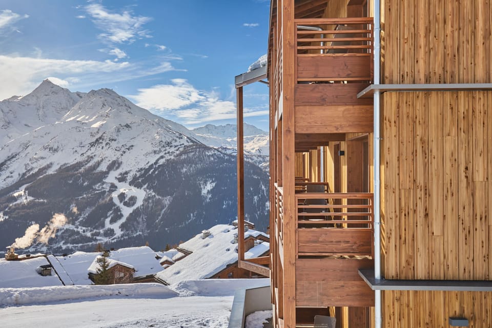 A wooden balcony with snow-laden rails overlooks the mountain slopes — the ski runs are steps away.