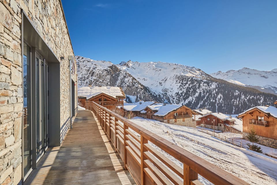Wooden walkway connecting the chalets, with snow-covered rooftops and alpine peaks framing the scene.