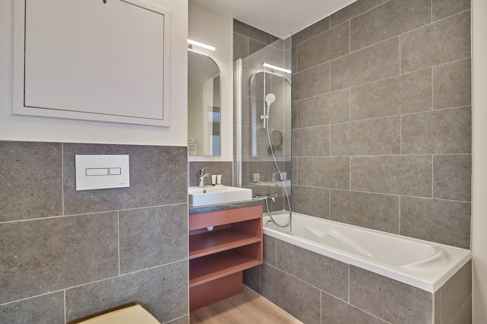 Grey-tiled bathroom with a bathtub, glass shower screen, and modern sink — towels included.