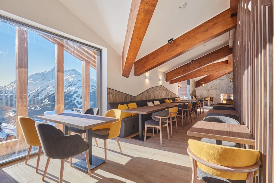 Dining area with a sloped wooden beam ceiling, grey and yellow cushioned seating, and mountain views through the windows.