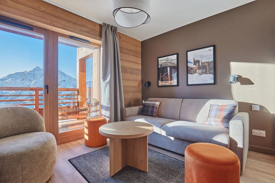 Living area with a grey sofa, plaid cushions, orange stools, wooden wall panelling, and a large window framing mountain views.