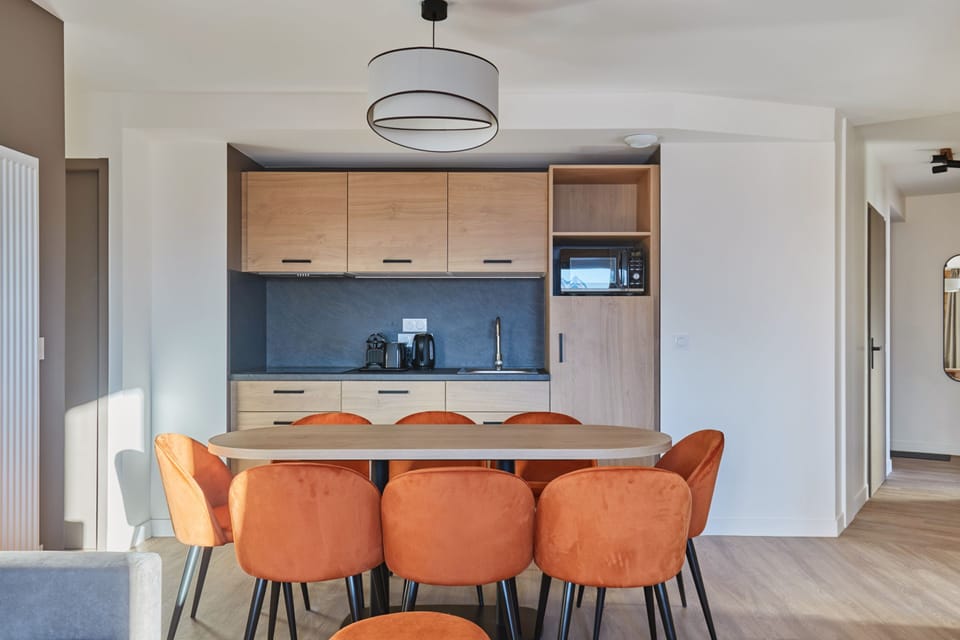 Kitchenette with light wood cabinets, black countertop, microwave, and eight orange chairs around the dining table.
