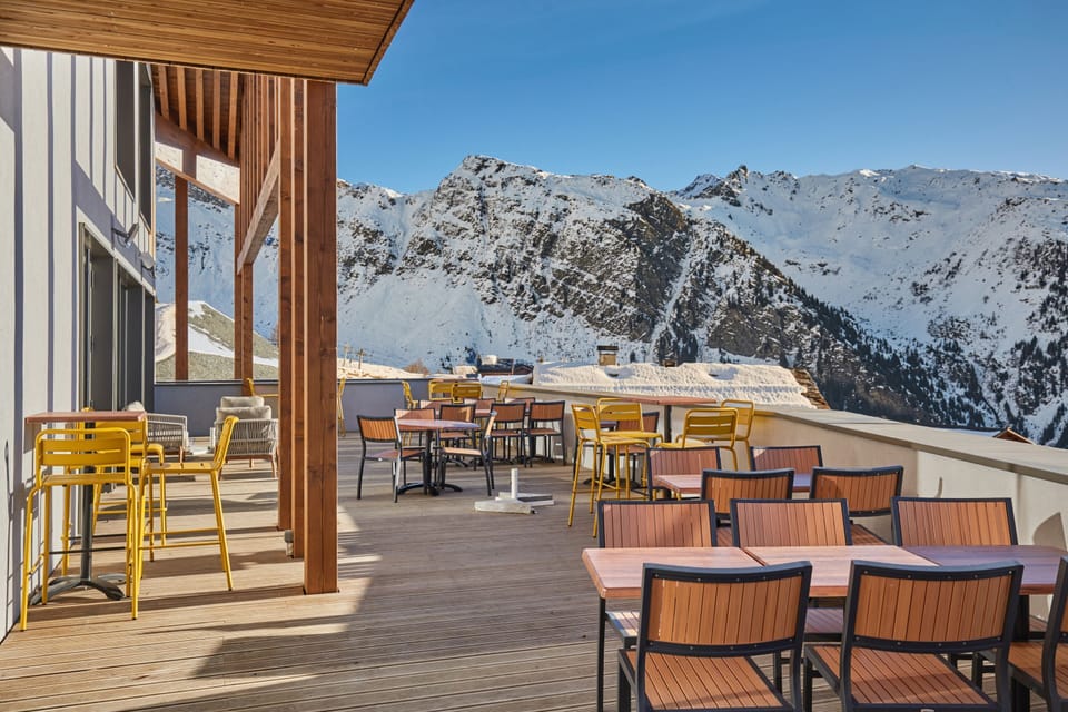 Outdoor terrace with yellow and black chairs around wooden tables, set against snow-capped peaks under a clear blue sky.