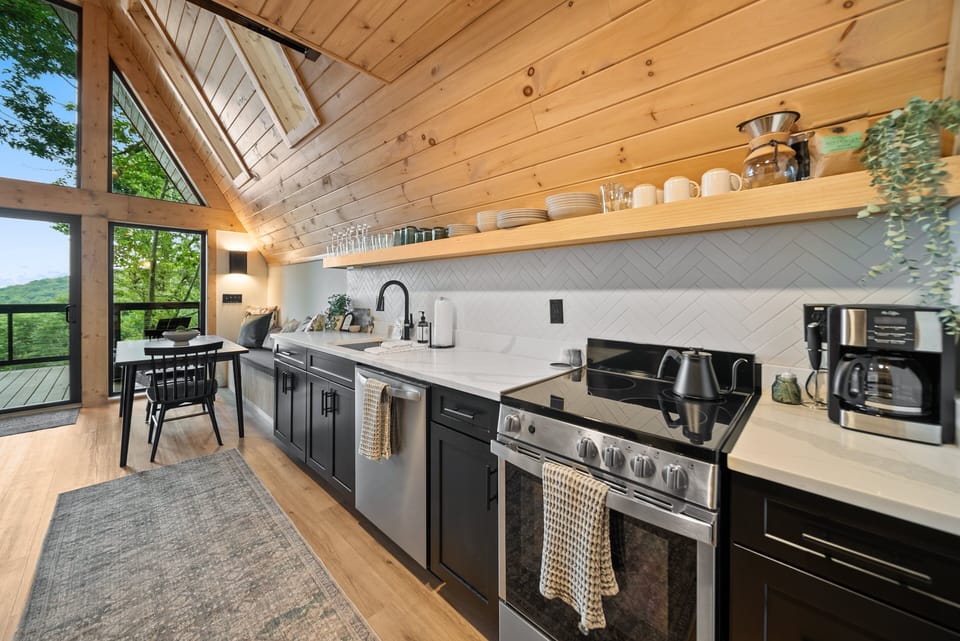 A bright, modern kitchen tucked under soaring wood-lined vaults, with treetop views just beyond the dining nook.