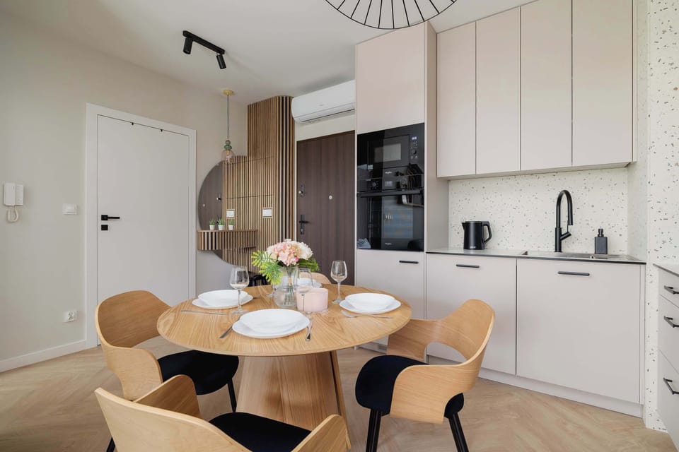A stylish dining room with a round wooden table, four comfortable chairs, elegant tableware and a decorative vase of flowers is adjacent to a modern kitchenette with semi-matt cashmere cabinets and black household appliances.