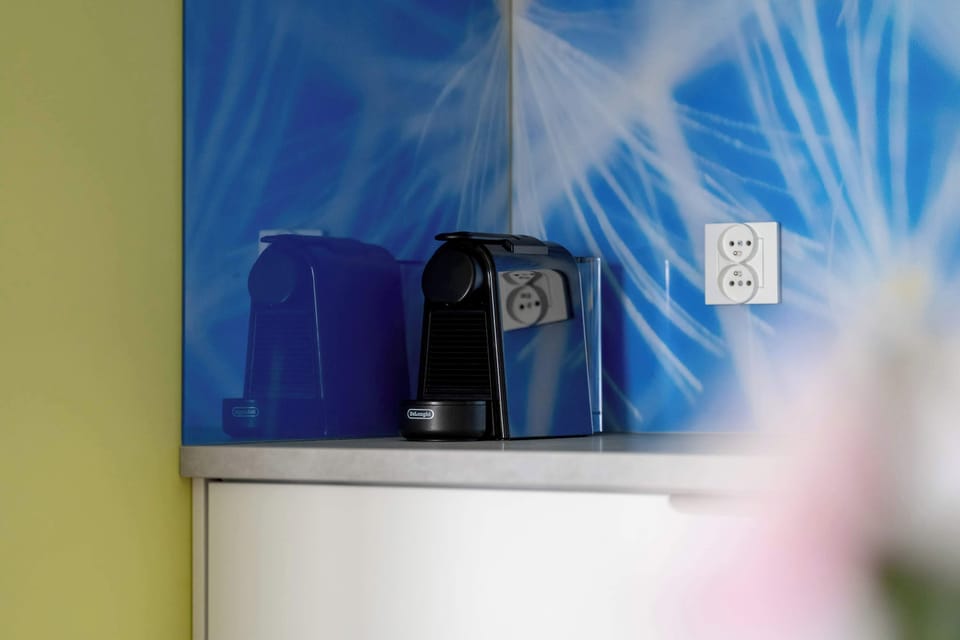 A modern kitchenette with a blue coffee maker, sleek countertop, and a decorative wall with white patterns creates a cozy space for preparing drinks.