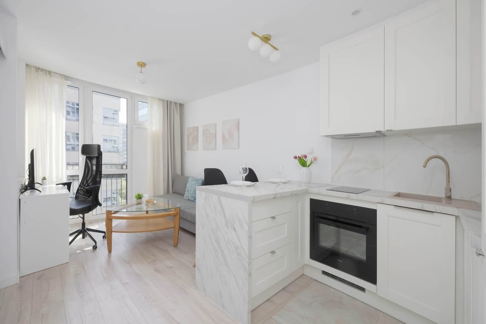 An open-plan view captures the seamless connection between the kitchen island and the living area workspace. The entire apartment is finished with light wood floors and crisp white walls, maximizing the sense of space.