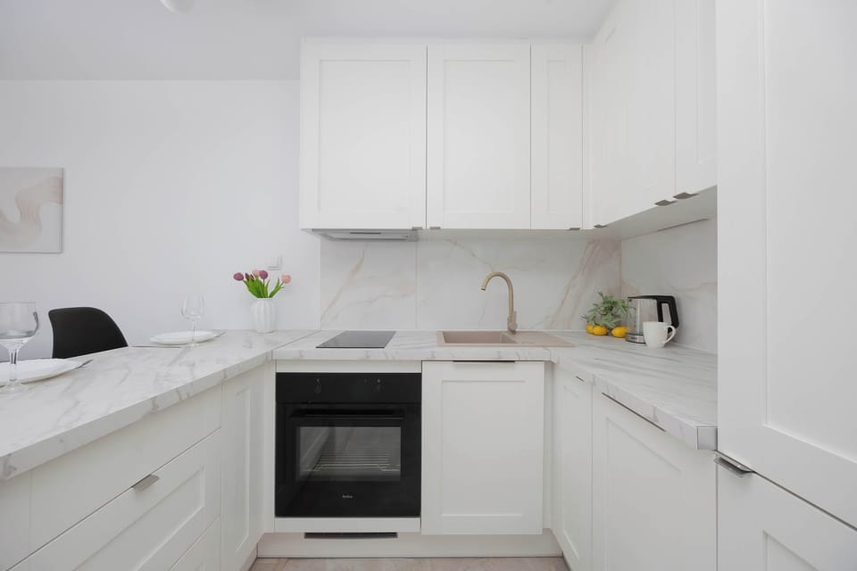A bright, white L-shaped kitchenette includes seamless cabinetry and a marble-patterned countertop with a matching backsplash. A built-in black oven and an electric stovetop create a clean, contemporary cooking area.
