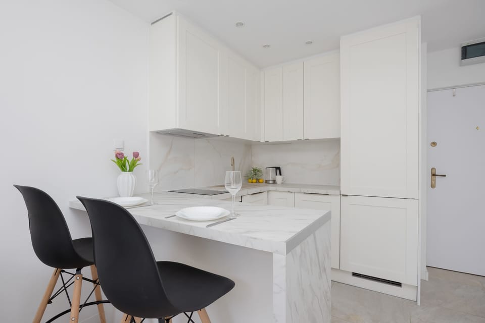 The kitchenette corner features a compact island bar paired with two black chairs with wooden legs. Tall white pantry cabinets line the back wall, extending all the way to the ceiling next to the apartment entryway.