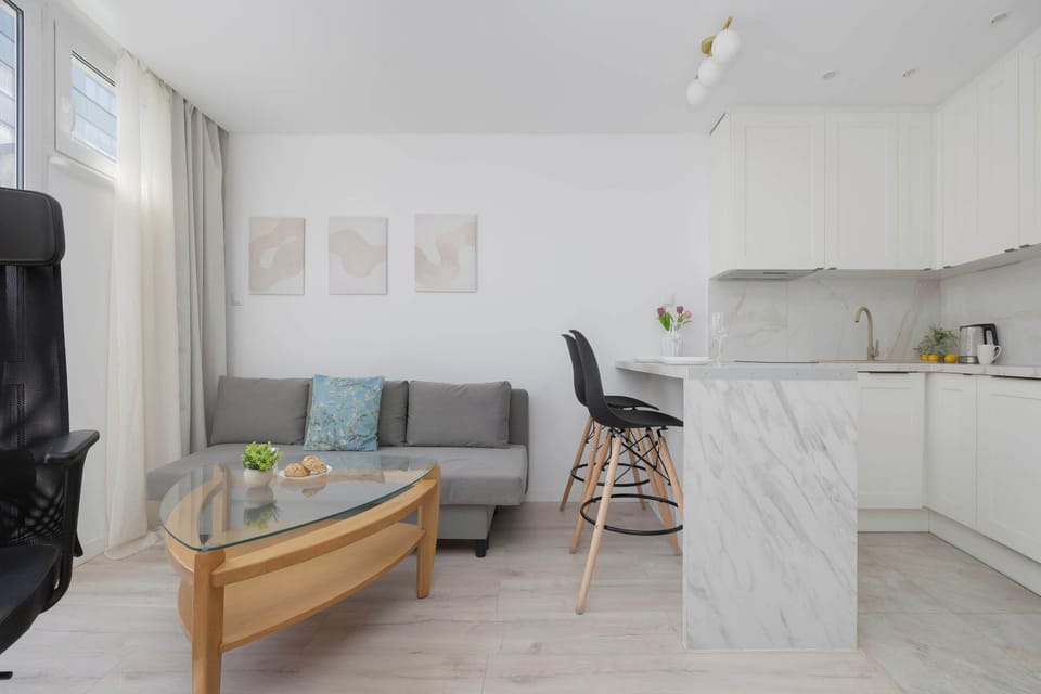 A broad shot of the studio space captures the entire living arrangement, from the grey sofa to the kitchen island. Black minimalist bar stools stand tucked under the marble-effect breakfast bar counter.