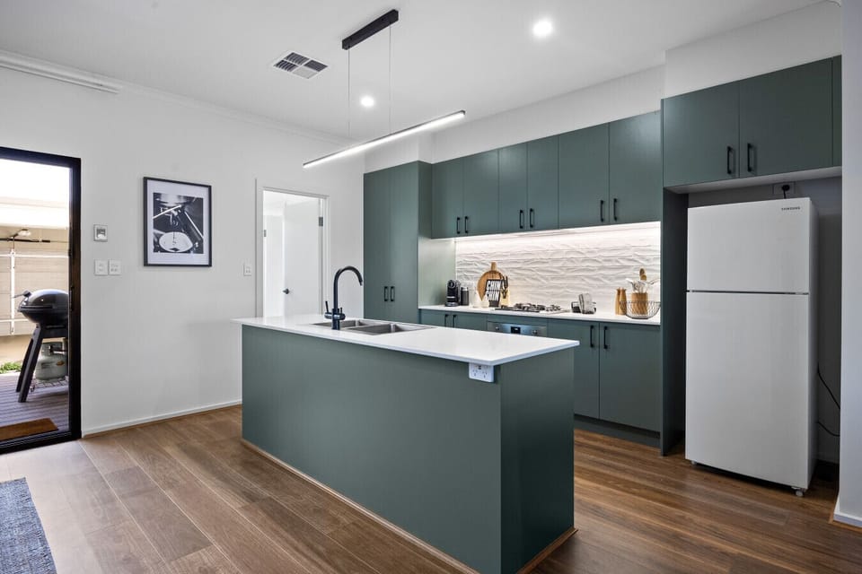 The kitchen combines deep green cabinetry and stone benchtops with a gas cooktop, oven, dishwasher, microwave, fridge, and pod coffee machine for effortless meal preparation.