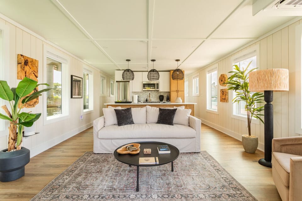 Sun-drenched main living area with a designer slipcovered sofa, woven rattan pendants, and 360° windows framing the marina — coastal luxury from every angle.
