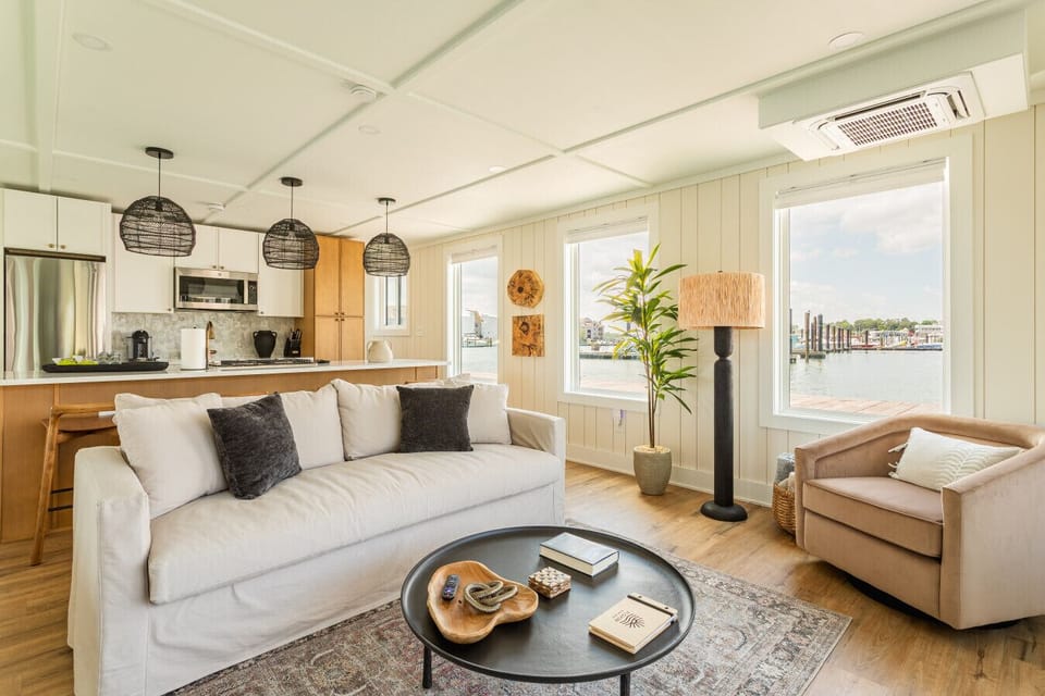 Bright coastal living room with a designer queen sleeper sofa, rattan floor lamp, and walls of windows showcasing the Virginia Beach Fishing Center marina.