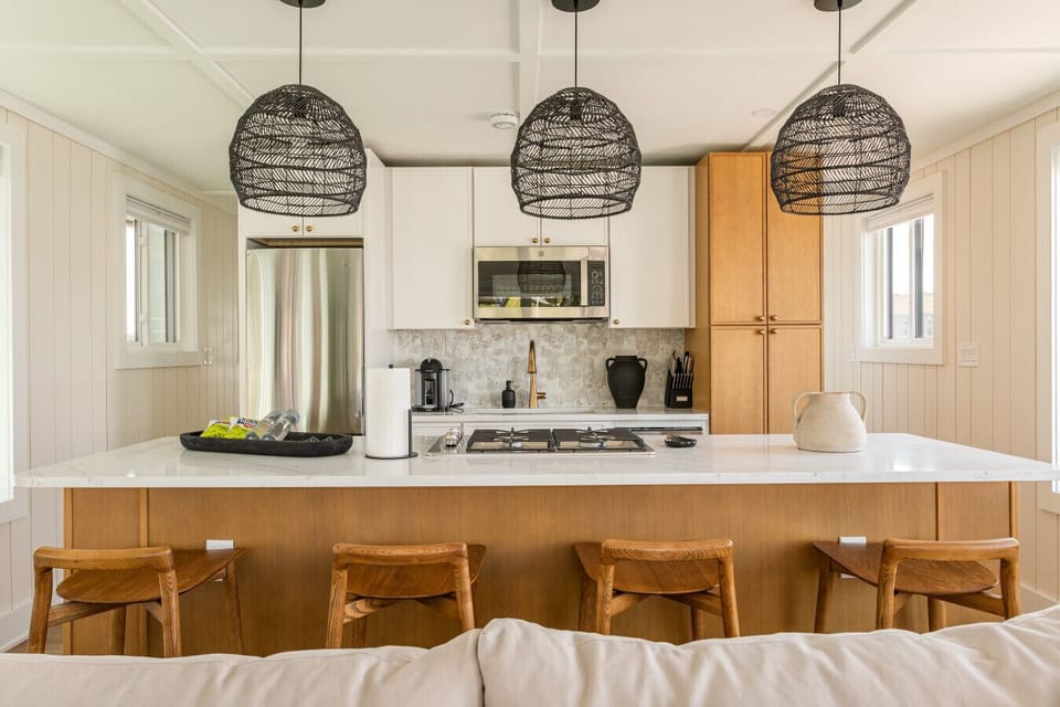Oversized quartz-topped kitchen island with four wood barstools, a four-burner gas cooktop, and woven rattan pendant lights — the airy heart of this coastal floating home.