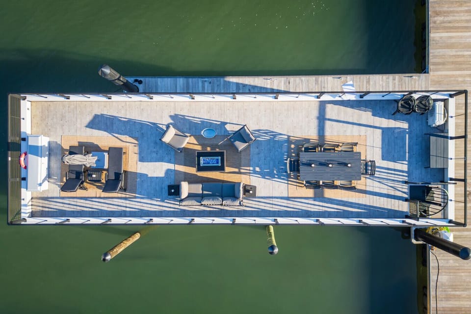 Top-down aerial of the private rooftop deck — eight-seat dining, lounge sectional, fire pit, and chaise loungers — floating above the green waters of Rudee Inlet.