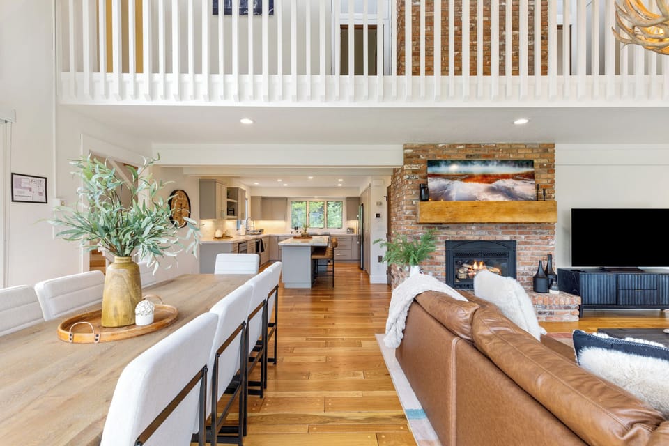 This open-concept living space invites you to relax by the brick fireplace or gather around the dining table. Warm wood floors flow seamlessly into the bright modern kitchen, while the loft above adds extra character and charm.