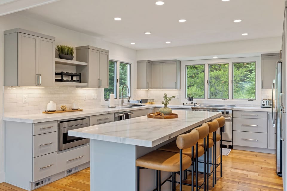 Bright, modern kitchen with sleek countertops, abundant workspace, and a spacious island. Natural light streams through large windows overlooking lush greenery, perfect for gathering and meal prep.