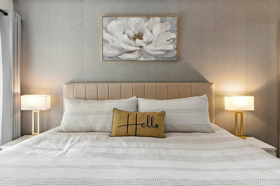 Experience restful nights in this beautifully styled bedroom featuring a plush king-size bed with crisp white linens and a charming "Hello" pillow. Soft bedside lighting and elegant floral artwork enhance the serene atmosphere.