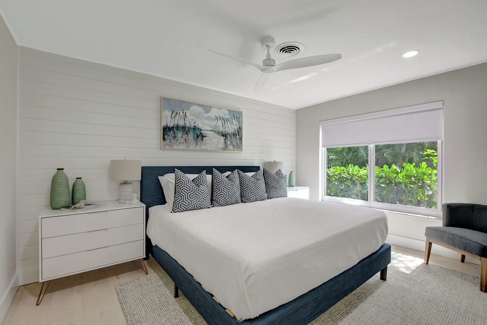 Settle into a comfortable bedroom with coastal styling, a blue headboard, ceiling fan, and a window overlooking greenery.