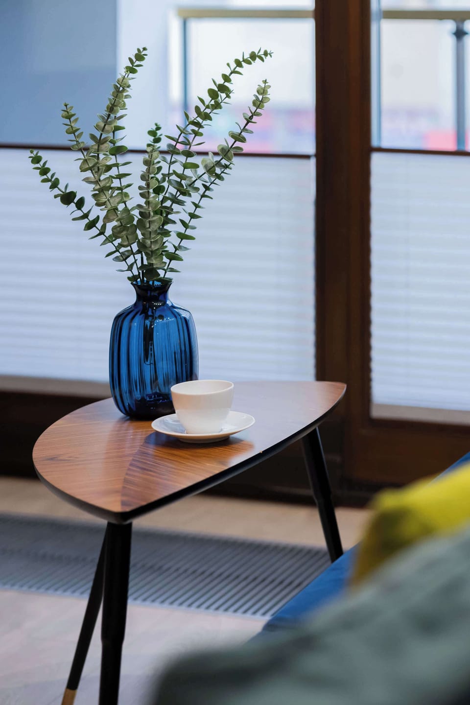 A small round wooden table with a blue vase and greenery, placed next to a large window with blinds. The corner looks peaceful and perfect for relaxing or reading.