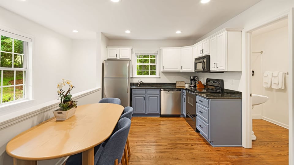 Full kitchen — white upper cabinets, blue-gray lowers, stainless steel appliances, 6-person dining table with a green view.