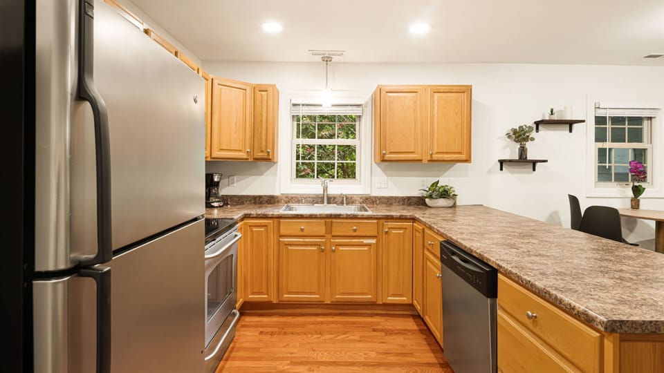 Scan the full kitchen run from this angle — full-size fridge, window above the sink with outdoor greenery, counter space that lets two people cook at the same time.