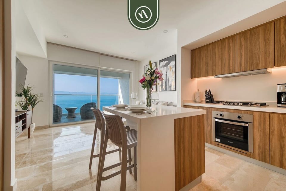 View across a modern white waterfall island set with dining chairs, looking out toward large balcony glass doors.