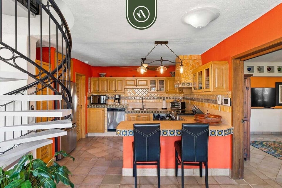 A spacious kitchen view featuring a tiled breakfast bar with leather barstools, set against a vibrant orange accent wall.