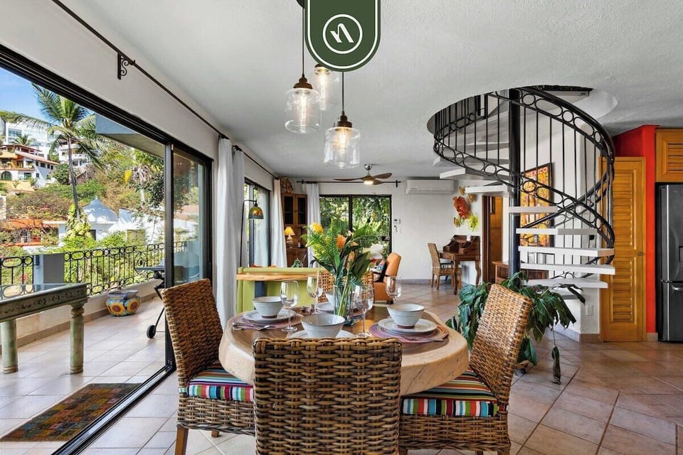 An elegant dining setup showing a round table with wicker chairs, situated next to a striking black spiral staircase.