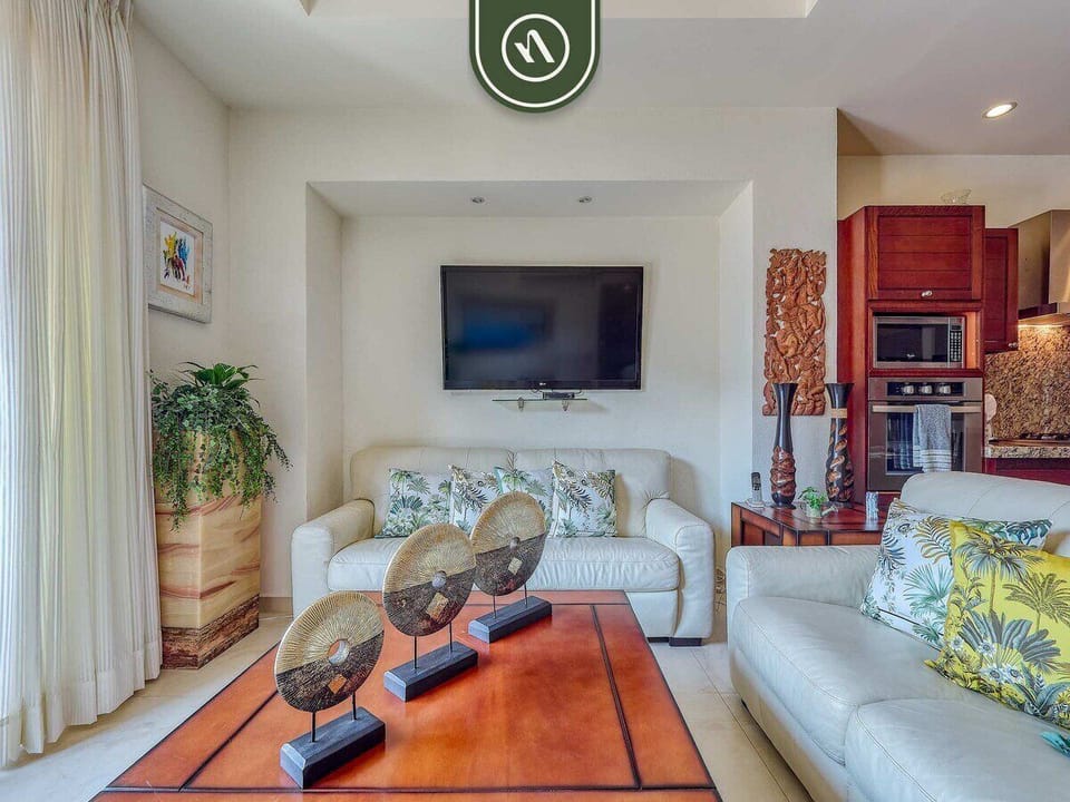 Unwind in this comfortable living room. It features plush white leather sofas, a flat-screen TV for entertainment, and tasteful decor, all seamlessly connected to the kitchen area.