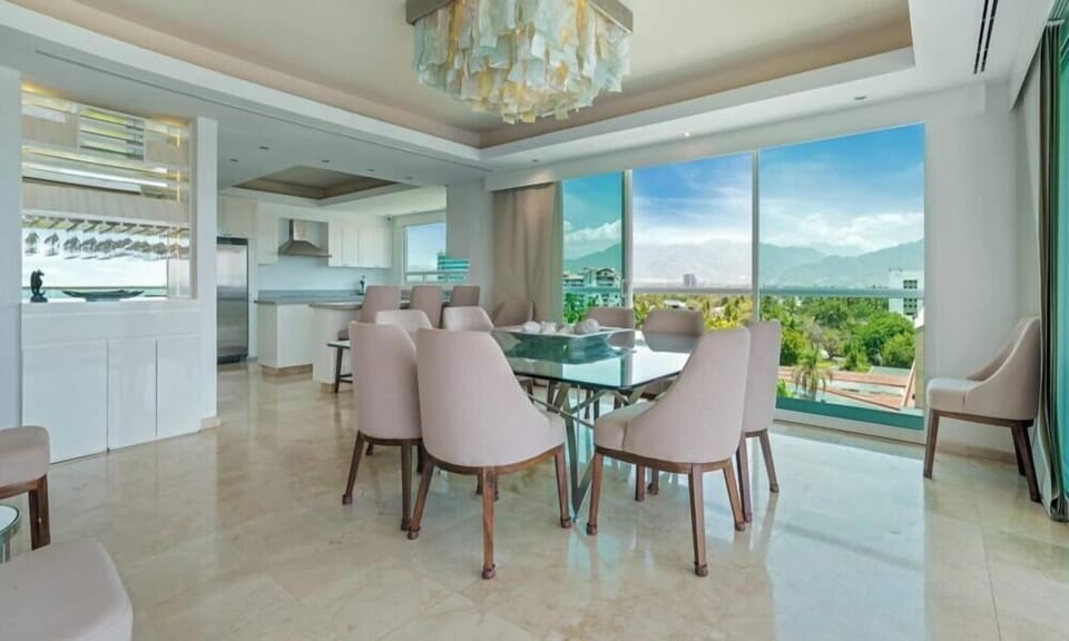 Elegant dining setup featuring a glass-top table, soft pink upholstered chairs, a unique artistic chandelier, and panoramic mountain views.