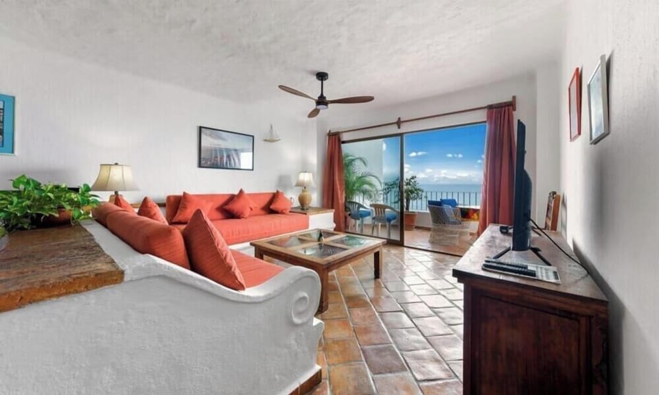 A comfortable sitting area featuring a red sofa, a flat-screen TV on a media console, and large glass doors showcasing ocean views.