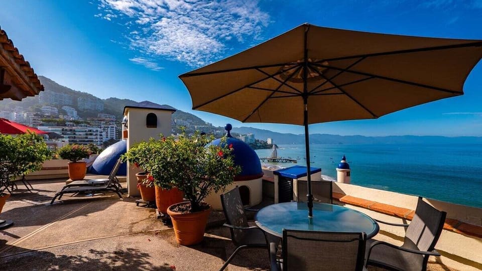 Shaded outdoor seating area with a patio umbrella, potted plants, and panoramic views of the coastal blue waters.