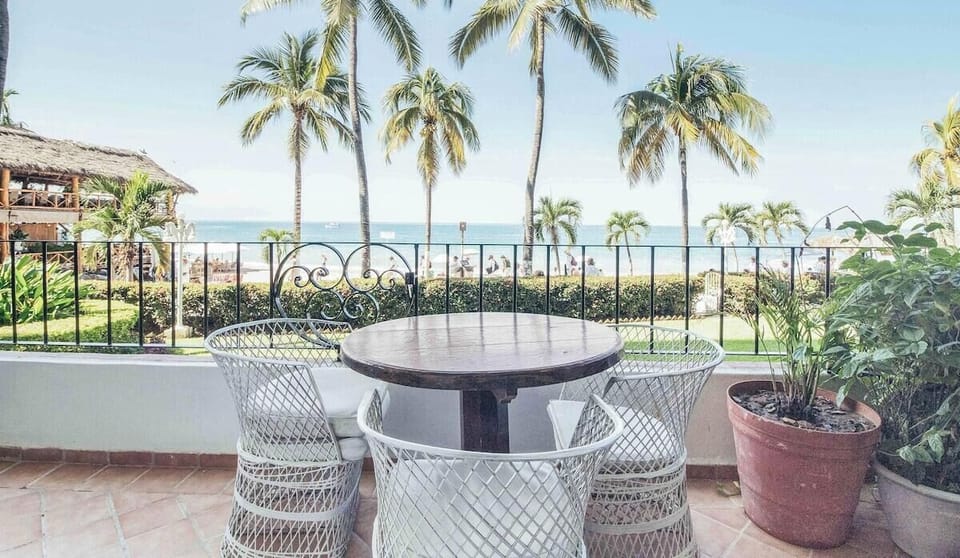 A private outdoor patio with woven chairs and a table, overlooking a sandy beach and palm trees.