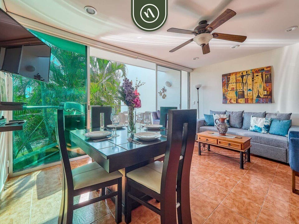 This wide shot showcases the spacious open-plan layout. The dining area flows effortlessly into the living room, with large glass doors providing a lush green backdrop and an abundance of natural light.