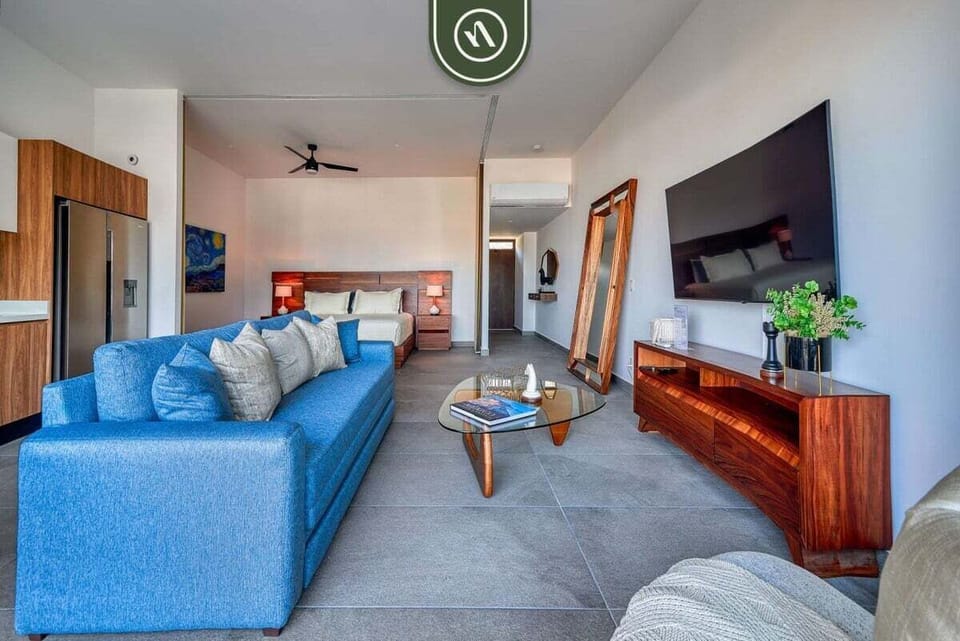 Modern living space featuring a bright blue sofa, a long wooden entertainment console with a large TV, and a view into the bedroom area.