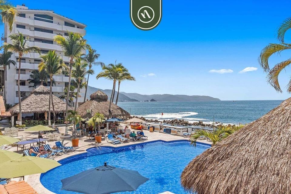 Sparkling blue swimming pool area bordered by palm trees and palapa structures right beside the ocean.