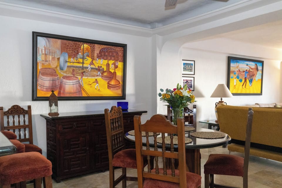 A cozy dining space featuring a round glass-topped table, traditional wooden chairs, a matching carved sideboard, and a vibrant yellow painting.