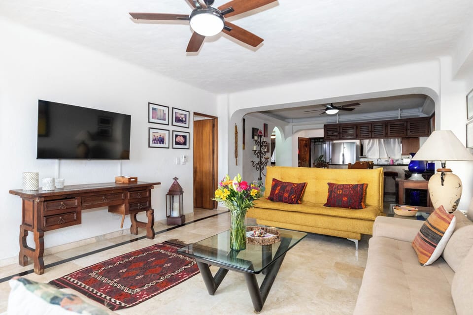 A spacious and comfortable living area featuring a mix of seating, a wall-mounted TV above a classic wooden console, and an arched entryway leading into the kitchen.