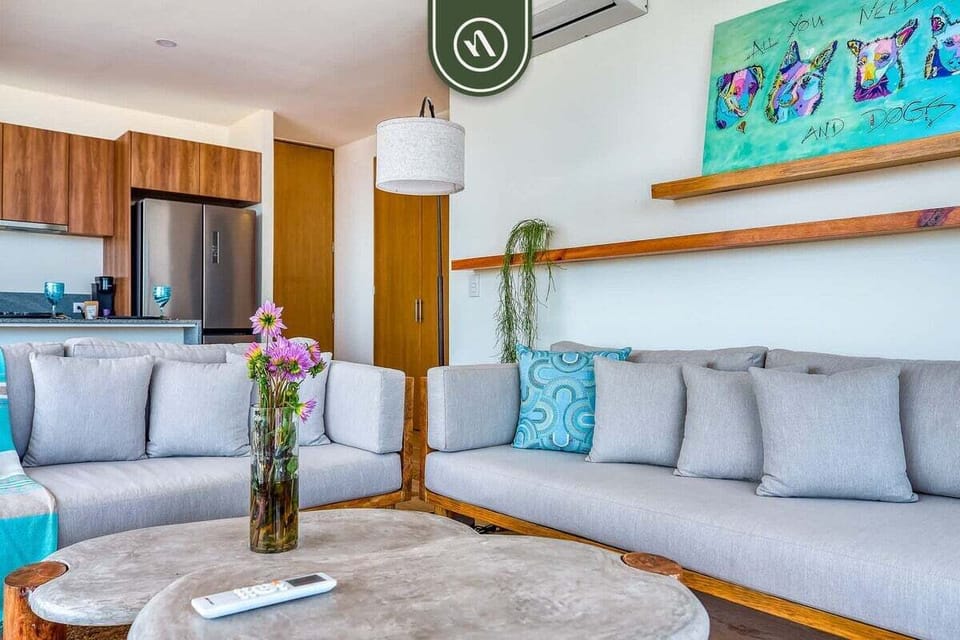 Unwind in a chic, airy living space boasting plush grey sectional sofas, natural wood elements, vibrant accent decor, and a seamless view into the adjacent kitchen.