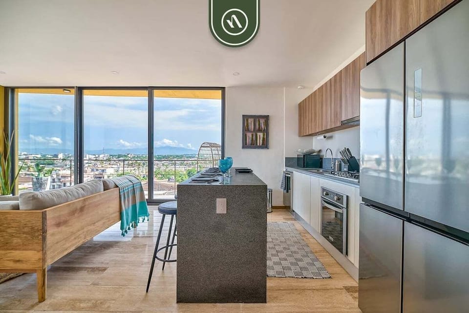 A detailed view of the culinary space highlighting the large stainless steel refrigerator, stone countertops, and the open transition into the bright living area.