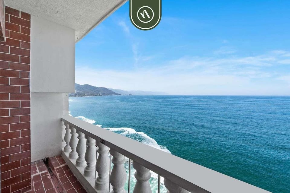 Looking out from a classic balustrade terrace across deep blue ocean waters toward rolling green hills.