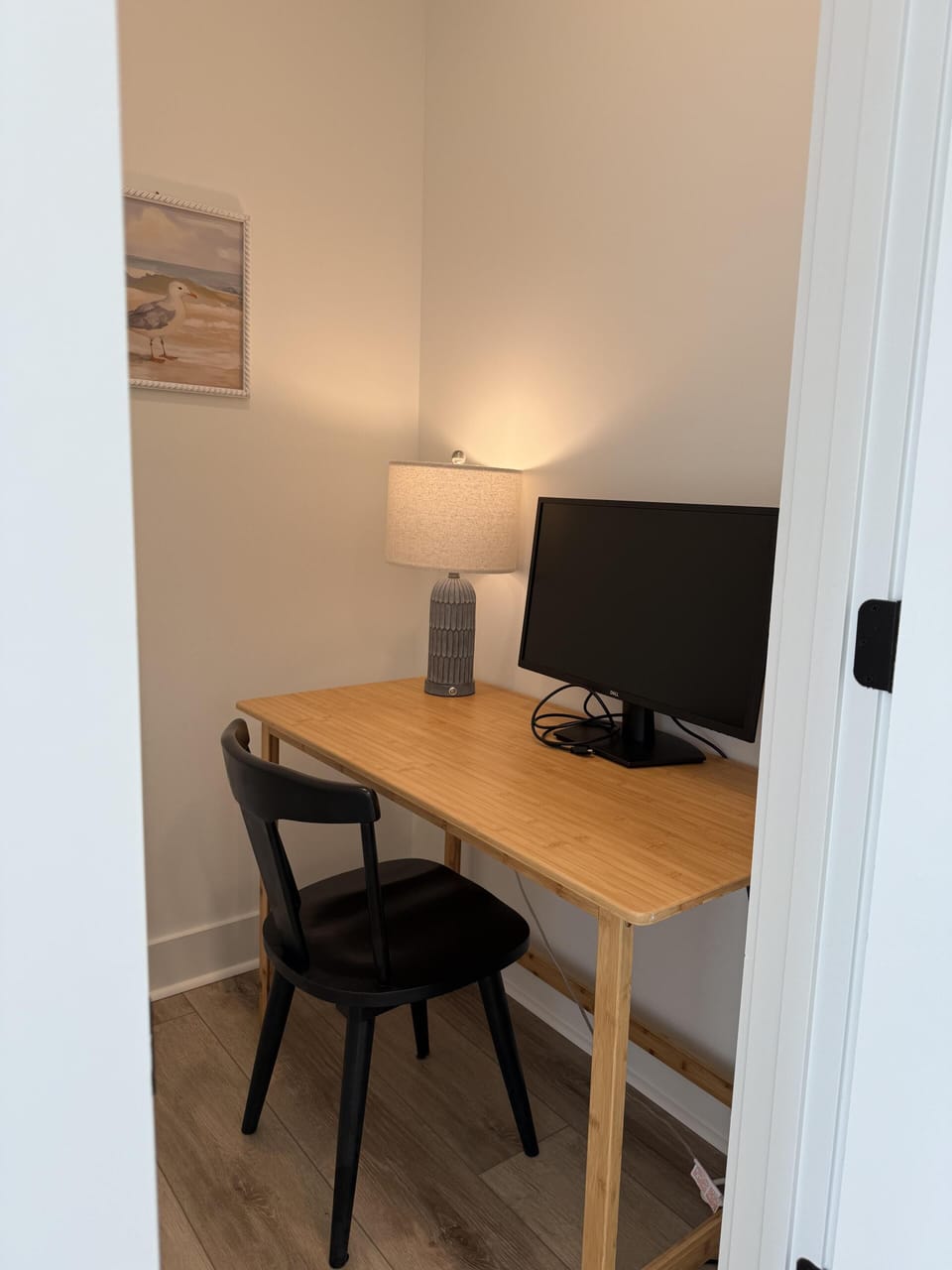 Small office nook, wood desk, chair, lamp, wall art, open door, nearby chair, window light, wood floors, bright coastal interior.